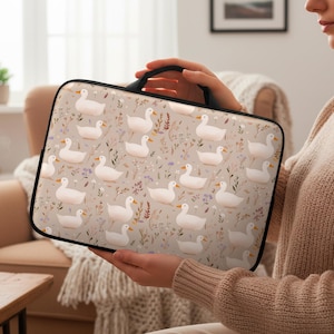 May include: A beige laptop sleeve featuring a repeating pattern of white ducks and floral accents. The sleeve has a black handle and trim. The design includes illustrations of ducks, leaves, and small purple flowers.
