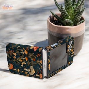 May include: A dark teal phone case featuring a mushroom and floral design. The case is open, displaying the phone's screen and the Samsung logo. The case has a hinged design and is decorated with orange, brown, and gold accents.