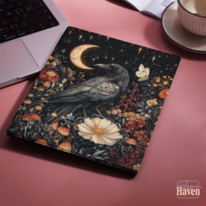 以下が含まれることがあります： 星空と三日月を背景に、大きな白い花にとまっているカラスの精巧なイラストが描かれた装飾的なラップトップスリーブ。デザインには、カラフルな花と蝶が含まれています。