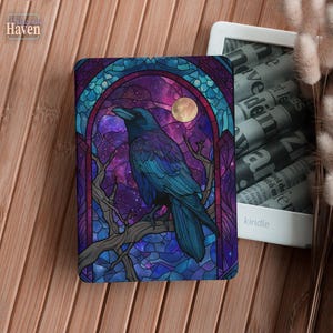 May include: A Kindle cover featuring a stained-glass design with a raven perched on a branch. The artwork includes a purple and blue night sky with a full moon. The cover is on a wooden surface.