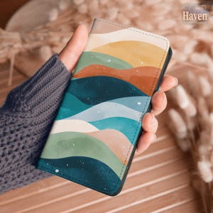May include: A rectangular phone case featuring a layered, wavy landscape pattern in shades of mustard yellow, burnt orange, teal, light blue, and sage green. The design includes subtle white speckles throughout, with black stitching along the edges.