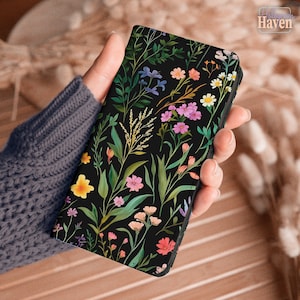 Könnte beinhalten: Eine schwarze Handyhülle mit einem lebendigen Blumenmuster. Das Design zeigt rosa, gelbe und weiße Blumen sowie grüne Blätter und Stiele. Die Hülle wird in der Hand einer Person gehalten.