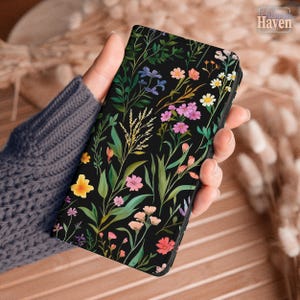 May include: A rectangular phone case featuring a black background with a vibrant botanical print of various wildflowers, including yellow, pink, purple, and white blossoms with green stems and leaves arranged in a scattered, natural pattern.