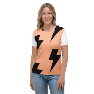 May include: A peach-coloured t-shirt featuring a black lightning bolt pattern. The sleeves are white. The shirt is worn by a person in blue jeans.