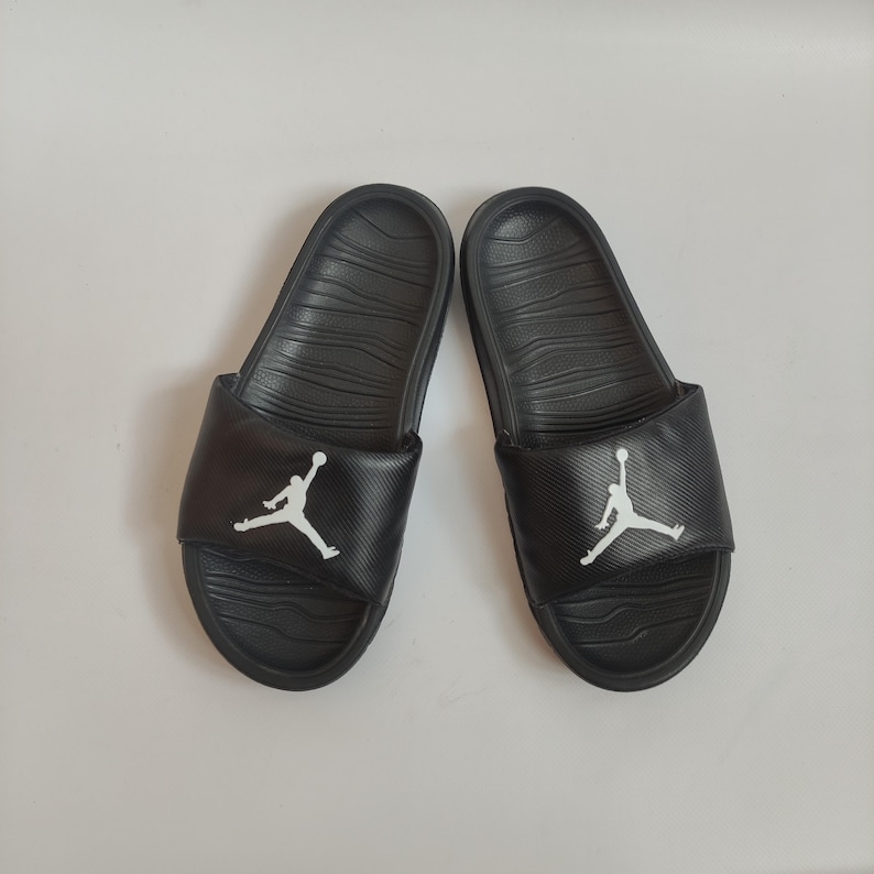 May include: Pair of black slides with a textured footbed and a wide strap. Each strap features a white Jumpman logo. The slides are designed for casual wear and are suitable for various activities.