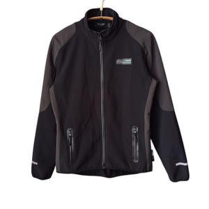 Puede incluir: Chaqueta negra y gris con cremallera y cuello alto. La chaqueta presenta un pequeño logotipo en el pecho y dos bolsillos con cremallera. Se ven franjas reflectantes en las mangas. La chaqueta está colgada en una percha.