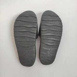 May include: A pair of black slide sandals, viewed from the bottom, with a textured sole design. The sandals feature the iconic Jumpman logo on the heel. The soles have a wavy pattern for grip and comfort.