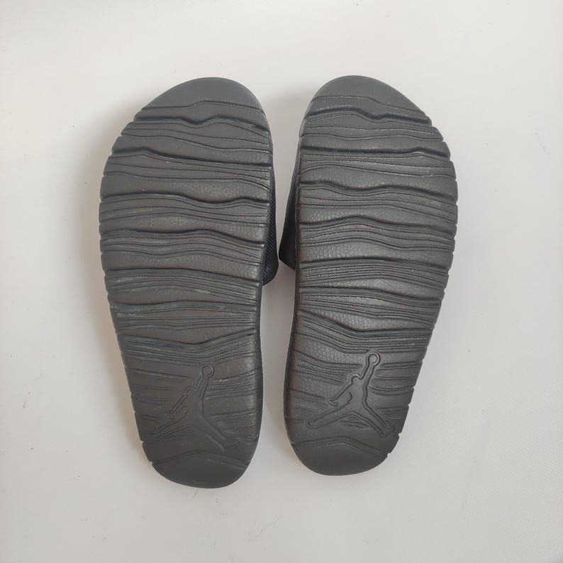 May include: A pair of black slide sandals, viewed from the bottom, with a textured sole design. The sandals feature the iconic Jumpman logo on the heel. The soles have a wavy pattern for grip and comfort.