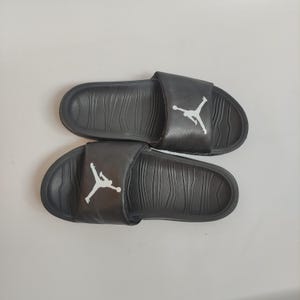 May include: A pair of black slides with a white Jumpman logo on the strap. The slides have a textured footbed and are designed for casual wear. The slides are shown on a white background.