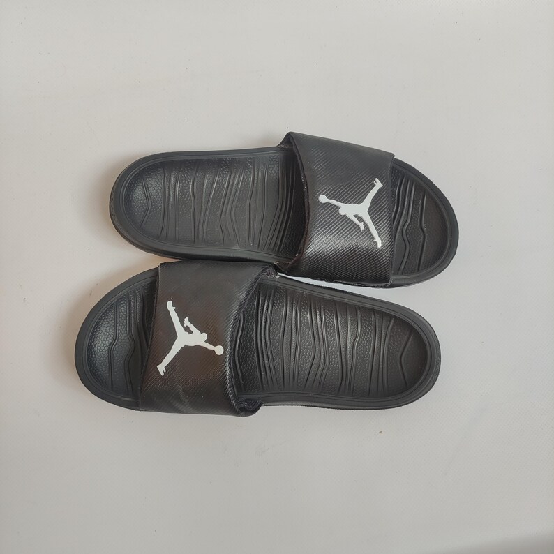 May include: A pair of black slides with a white Jumpman logo on the strap. The slides have a textured footbed and are designed for casual wear. The slides are shown on a white background.
