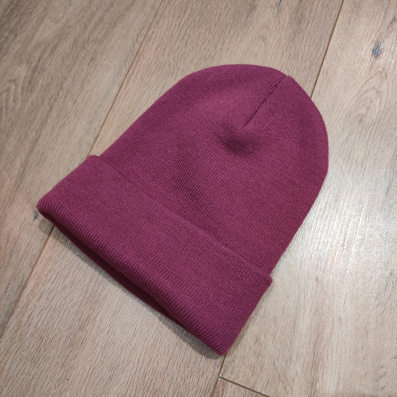 Puede incluir: Un gorro burdeos liso con un borde doblado. El gorro est&aacute; hecho de un material suave y tejido y est&aacute; sobre una superficie de madera. El gorro es de un tono rojo-p&uacute;rpura intenso.
