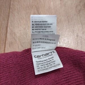Puede incluir: Primer plano de una tela de punto burdeos con varias etiquetas adjuntas. Las etiquetas incluyen instrucciones de lavado en varios idiomas, texto en japon&eacute;s y el logotipo de Carhartt Work In Progress. Las etiquetas tambi&eacute;n incluyen la direcci&oacute;n del fabricante.