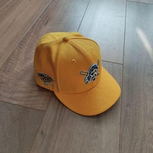 Op de afbeelding: Een mosterdgele baseballcap met een platte klep. De cap heeft een logo van een schedel en gekruiste botten met een honkbalknuppel en de tekst "ATLANTA BRAVES".