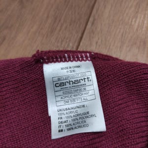 Puede incluir: Primer plano de un gorro de reloj acr&iacute;lico burdeos con una etiqueta. La etiqueta dice "Carhartt" y "Acrylic Watch Hat". El sombrero es de talla &uacute;nica.