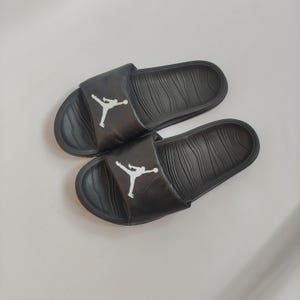 May include: Pair of black slides with a textured footbed and a white Jumpman logo on the strap. The slides are made of a durable material and are designed for comfort and style.