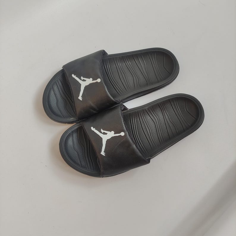 May include: Pair of black slides with a textured footbed and a white Jumpman logo on the strap. The slides are made of a durable material and are designed for comfort and style.
