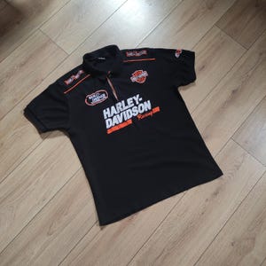 Könnte beinhalten: Schwarzes Poloshirt mit orangefarbenen und weißen Harley-Davidson Racing-Grafiken. Das Shirt hat einen Kragen, kurze Ärmel und mehrere Harley-Davidson-Logos. Das Shirt besteht aus einem leichten Material.