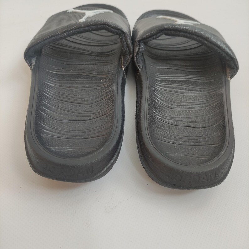 May include: A pair of black Jordan slides. Each slide features a molded footbed with a textured pattern and a wide strap with the Jordan logo in white. The word "JORDAN" is embossed on the sole.