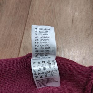 Puede incluir: Primer plano de una prenda de punto burdeos con una etiqueta blanca. La etiqueta muestra las instrucciones de lavado y la composici&oacute;n del tejido en varios idiomas, incluido el espa&ntilde;ol, e indica que la prenda es 100% acr&iacute;lico.