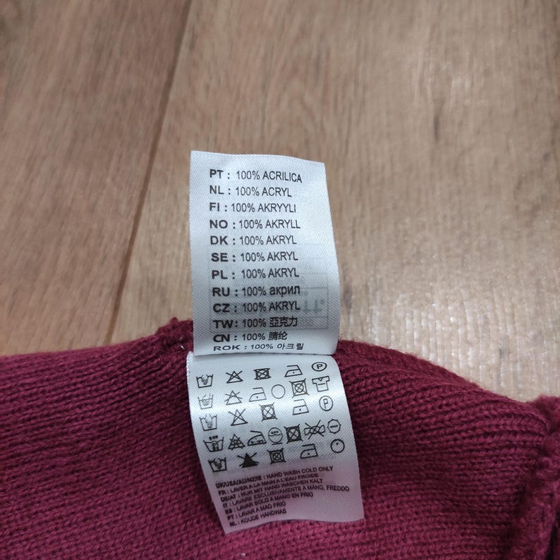 Puede incluir: Primer plano de una prenda de punto burdeos con una etiqueta blanca. La etiqueta muestra las instrucciones de lavado y la composici&oacute;n del tejido en varios idiomas, incluido el espa&ntilde;ol, e indica que la prenda es 100% acr&iacute;lico.