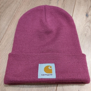 Puede incluir: Un gorro de punto magenta con un borde doblado. El gorro presenta un parche cuadrado con el logotipo de Carhartt, una 'C' dorada dentro de un cuadrado blanco y el nombre de la marca en texto blanco.
