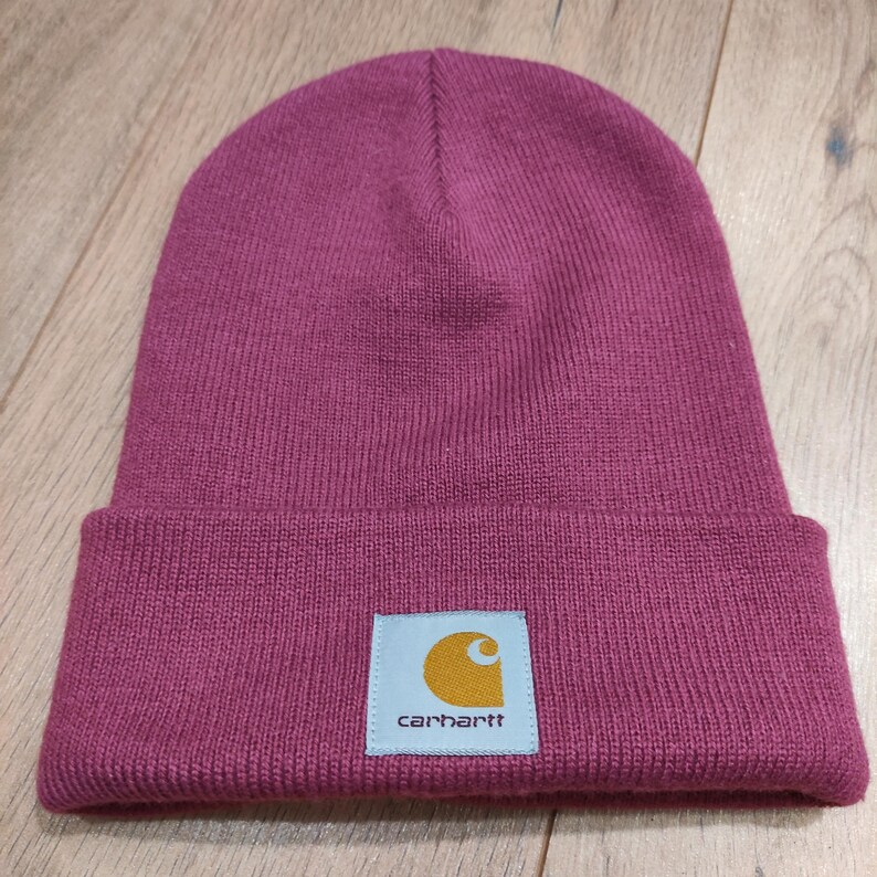 Puede incluir: Un gorro de punto magenta con un borde doblado. El gorro presenta un parche cuadrado con el logotipo de Carhartt, una 'C' dorada dentro de un cuadrado blanco y el nombre de la marca en texto blanco.