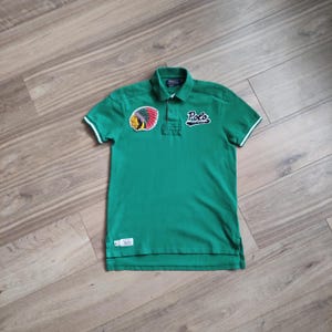 May include: Green polo shirt with white trim on the sleeves and collar. Features a patch with a Native American headdress and the word "Polo" in white script. A small tag is visible near the hem.