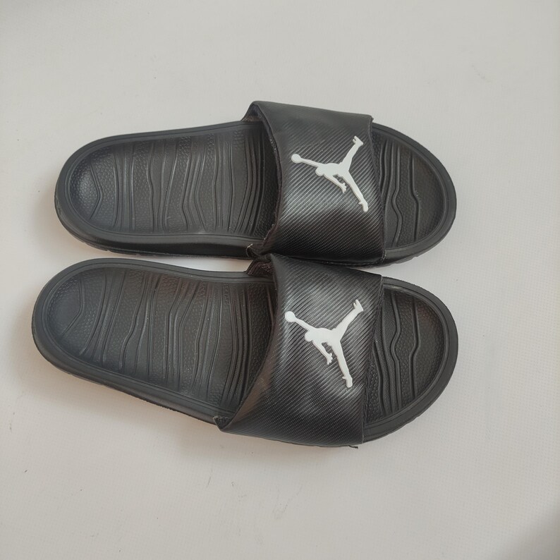 May include: Pair of black slide sandals with a textured footbed and a wide strap. The strap features a white Jumpman logo. The sandals are designed for casual wear and are suitable for various activities.