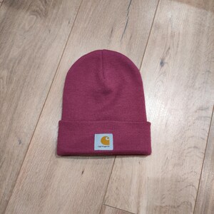 Puede incluir: Un gorro de punto burdeos con un borde doblado. El gorro presenta un peque&ntilde;o parche rectangular con el logotipo de Carhartt en amarillo y blanco. El sombrero est&aacute; hecho de un material suave y tejido.
