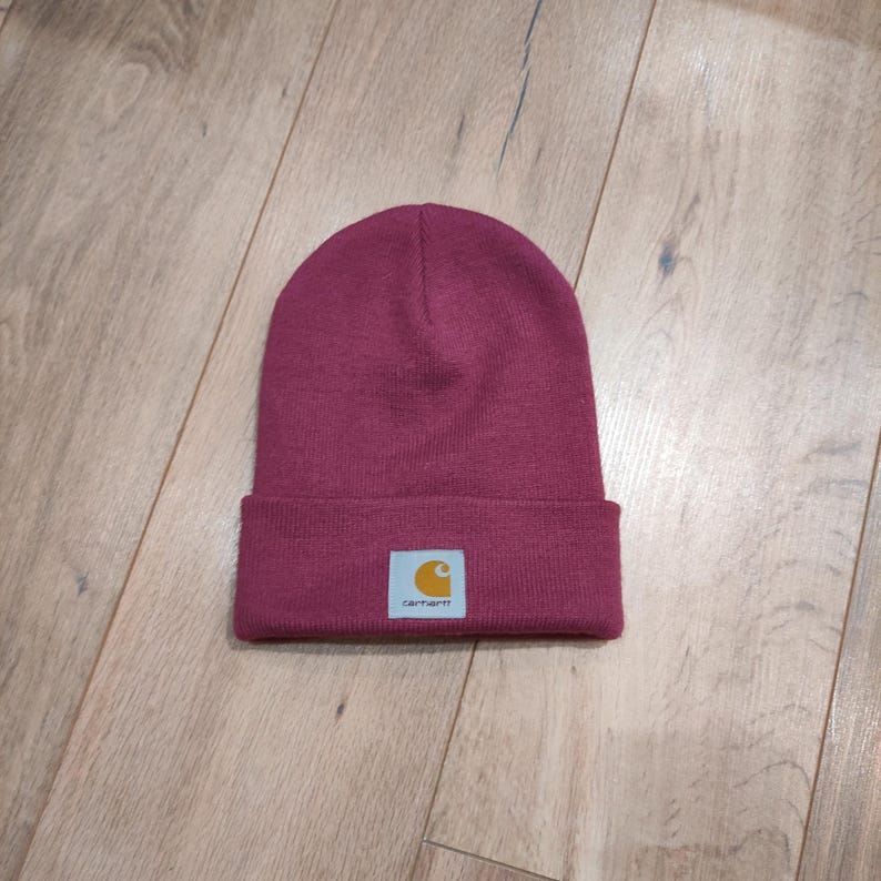 Puede incluir: Un gorro de punto burdeos con un borde doblado. El gorro presenta un peque&ntilde;o parche rectangular con el logotipo de Carhartt en amarillo y blanco. El sombrero est&aacute; hecho de un material suave y tejido.