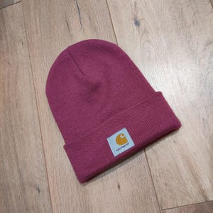 Puede incluir: Un gorro de punto burdeos con borde doblado. El gorro presenta una peque&ntilde;a etiqueta rectangular con el logotipo de Carhartt en amarillo y blanco. El gorro est&aacute; hecho de un material de punto acanalado.