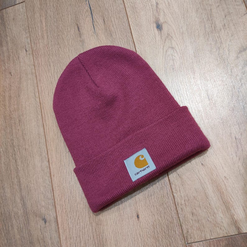 Puede incluir: Un gorro de punto burdeos con borde doblado. El gorro presenta una peque&ntilde;a etiqueta rectangular con el logotipo de Carhartt en amarillo y blanco. El gorro est&aacute; hecho de un material de punto acanalado.