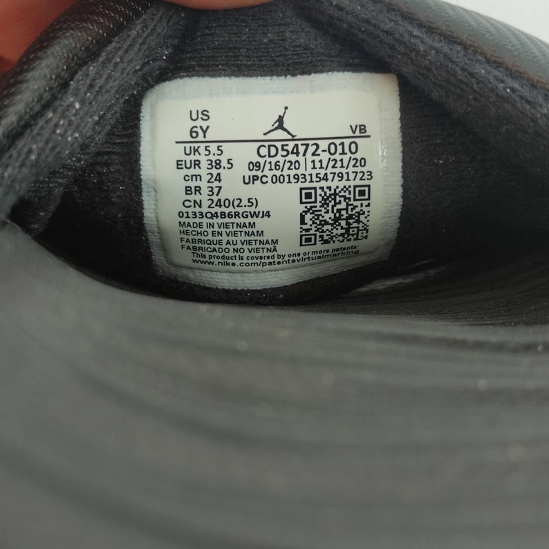 May include: Close-up of a black shoe label with white text. The label includes size information: US 6Y, UK 5.5, EUR 38.5, cm 24, and BR 37. The label also displays a product code, manufacturing details, and a QR code.