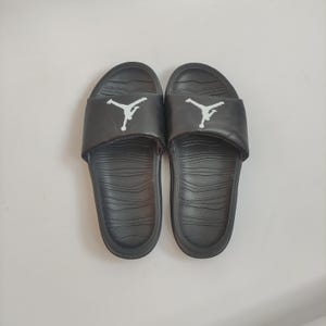 May include: Pair of black slide sandals with a white Jumpman logo on the strap. The sandals have a textured footbed and a solid black sole. These slides are suitable for casual wear and outdoor activities.