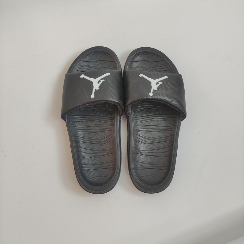 May include: Pair of black slide sandals with a white Jumpman logo on the strap. The sandals have a textured footbed and a solid black sole. These slides are suitable for casual wear and outdoor activities.