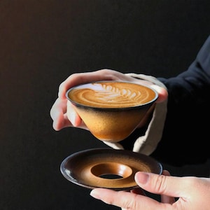 May include: A ceramic coffee cup and saucer set. The cup is filled with latte art, featuring a leaf design. The cup and saucer have a gradient color scheme, transitioning from a dark brown to a golden hue. The set is held in hands against a dark background.