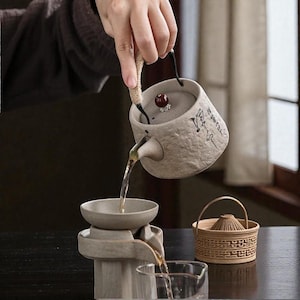 May include: A gray ceramic teapot with a woven handle and a small red knob on the lid is pouring liquid into a gray ceramic tea set. The set includes a small bowl and a glass. A small woven basket sits to the right.