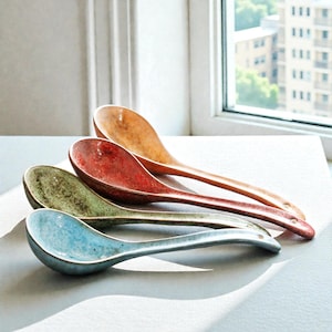 Może przedstawiać: Cztery ceramiczne łyżki w rzędzie, każda w unikalnym kolorze. Łyżki są w odcieniach niebieskiego, zielonego, czerwonego i pomarańczowego. Łyżki mają długi uchwyt i zaokrągloną miseczkę. Łyżki leżą na białej powierzchni w pobliżu okna.
