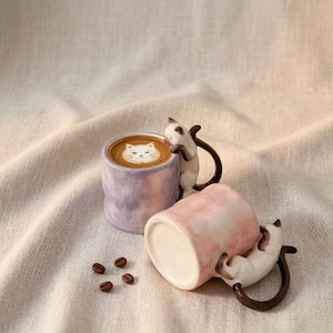 May include: Two ceramic coffee mugs with cat designs. One mug is upright, filled with coffee and a cat face design in the foam. The other mug is tipped over. Both mugs have a cat figurine attached. Coffee beans are scattered around.
