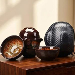 Puede incluir: Un juego de té de cerámica marrón con una tetera, dos tazas de té y un cuenco pequeño. Las tazas de té tienen un diseño interior único con una mezcla de tonos naranja, amarillo y marrón. También se incluye un estuche negro.