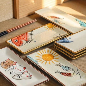 Peut inclure: Ensemble de plats de service rectangulaires en céramique avec des motifs peints à la main. Les plats présentent des illustrations de poissons, de soleils, de nuages et de chats. Chaque plat a une bordure dorée ou marron. Une paire de baguettes est posée sur le côté.