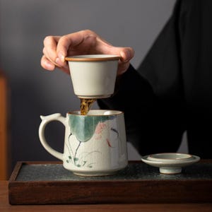 Puede incluir: Un juego de té de cerámica con un diseño floral. El juego incluye una taza con asa, un colador y una tapa. La taza está decorada con motivos florales verdes y rosas. Se está vertiendo té del colador a la taza.
