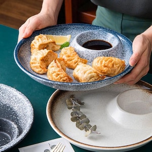 May include: A blue and white speckled ceramic serving plate with a built-in sauce bowl, holding fried dumplings and a lemon wedge. The plate is held by hands, with a second plate visible below.