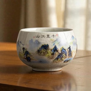May include: A white ceramic teacup with a landscape painting of blue mountains and green foliage. The cup has a crackle glaze and a rounded shape. The text in the image is in a foreign language.