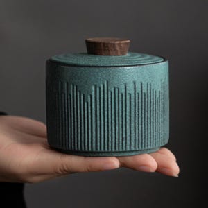 May include: A teal ceramic jar with a wooden lid. The jar has a cylindrical shape with vertical line detailing around the body. The lid is a dark brown, flat disc shape. The jar is held in a hand.