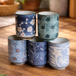 May include: A stack of five ceramic tea cups with floral patterns in shades of blue and white. The cups have a cylindrical shape with a gold rim and are arranged on a wooden surface.