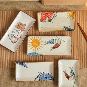 Puede incluir: Colección de platos rectangulares de cerámica con diseños pintados a mano. Los diseños incluyen gatos, peces, soles y nubes en una variedad de colores. Los platos tienen un fondo crema con ribetes marrones y dorados.