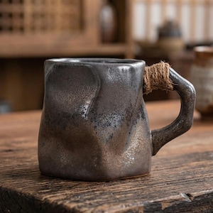 Puede incluir: Taza de cerámica gris oscuro con superficie texturizada y asa envuelta en cuerda. La taza tiene una base cuadrada y una forma irregular. Ideal para té o café. Dimensiones: aproximadamente 10 cm de altura.