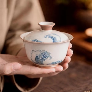 May include: A white ceramic tea bowl with a lid, featuring blue wave patterns. The rim and the lid's handle are a light brown color. The bowl is held in someone's hands.