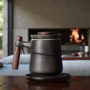 Puede incluir: Una taza de cerámica negra con asa y tapa de madera, sobre un posavasos negro. La taza tiene una superficie texturizada y un diseño único. El asa y la tapa son de madera. La taza está diseñada para té o café.